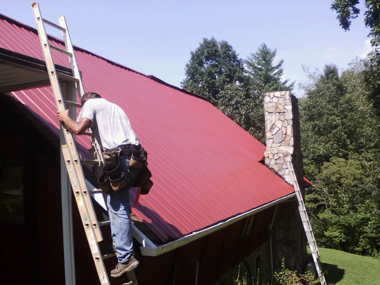 Roofer on a ladder beside a red metal roof and stone chimney — Soffit & Fascia Repair by Roofing Brothers in Bairdford